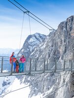 Zwei Erwachsene und zwei Kinder stehen auf einer Hängebrücke und unten sieht man verschneite Berge | © Josh Absenger