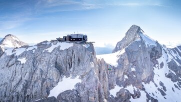 Dachstein Gletscherbahn fährt zwischen schneebedeckten Felsgipfeln unter blauem Himmel. | © Josh Absenger