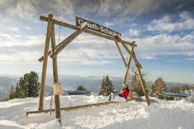 Person in red jacket on a wooden swing with alpine mountain view in the background. | © Lorenz Masser