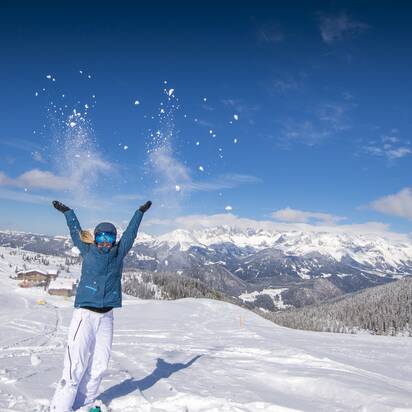 Woman throws snow in the air, smiling, with mountains and snowy valley in background. | © Lorenz Masser