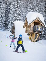 Zwei Kinder beim Skifahren vor verschneiten Bäumen und einer Hütte im Galstiland Galsterberg. | © Mirja Geh