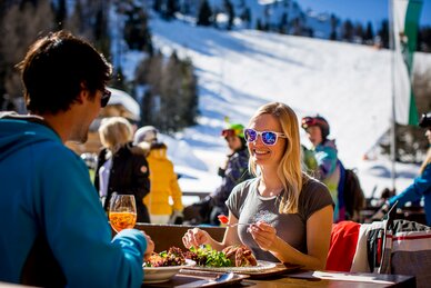 Woman with sunglasses enjoying food at the ski hut in sunshine, skiers in the background. | © Tom Lamm