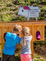 Two kids feel into a wooden tactile station with fabrics; a sheep sign presents a riddle on the themed mountain trail. | © Hauser Kaibling