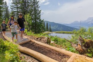Family with kids walking barefoot on a trail of gravel and soil at Hauser Kaibling, with mountain lake and Alps in the background. | © René Eduard Perhab