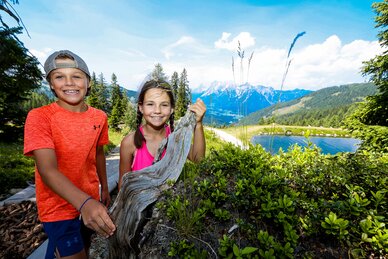 Two smiling kids by a tree stump at Schafsinn pond with mountain views in summer. | © Stefan Pajman