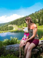 Woman and child sit on a log by a clear mountain lake, looking at the water, surrounded by forest and a hiking path. | © Stefan Pajman