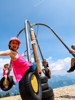 Kids swinging on a tire carousel with mountain view, laughing and stretching arms happily toward the camera. | © Stefan Pajman