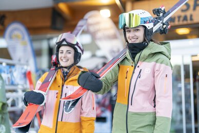 Two skiers in colorful ski outfits with skis in front of ski rental, ready for skiing. | © Bernhard Moser