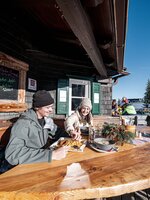 Two people eat outside at a ski hut with wooden bench and snowy background. | © Hauser Kaibling