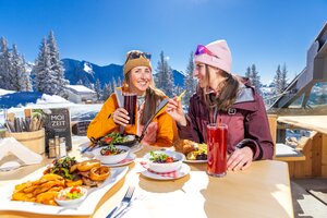 Two women eat and drink in the sun on the terrace with snowy mountain views. | © Hauser Kaibling