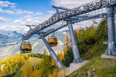 Yellow Hochwurzen gondolas move through autumn forest with scenic views of alpine valley below. | © Alexander Klünsner