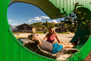 Hiking couple relaxes on heart-shaped bench with view of alpine huts and sunny mountain scenery. | © Christine Höflehner