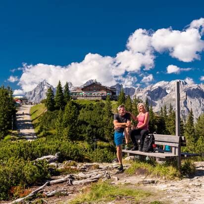 Hiking couple rests on bench overlooking Hochwurzen hut and rugged mountain peaks in bright sunshine. | © Christine Höflehner