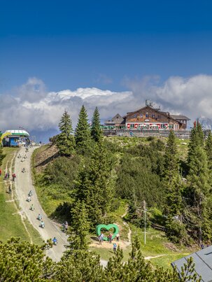 Go-kart riders and hikers at Hochwurzen summit with alpine hut, heart-shaped spot and mountain view. | © Planai/Kovacsics