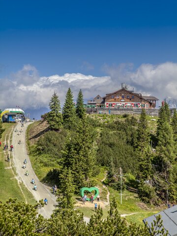 GoKart-Fahrer und Wanderer am Gipfel der Hochwurzen mit Almhütte, Herzplatz und Panorama unter blauem Himmel. | © Planai/Kovacsics