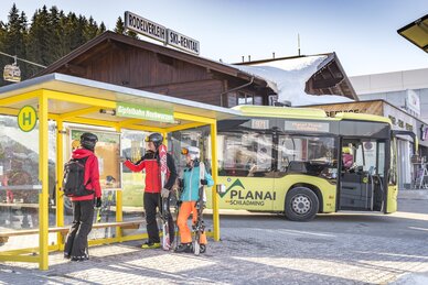 Skiers with equipment at Hochwurzen ski bus stop, ski bus in background. | © David Stocker