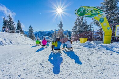 Vier Rodler starten am Hochwurzen-Rodelweg, sonniger Wintertag mit Bergen im Hintergrund. | © René Eduard Perhab