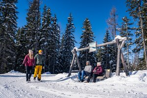 Winter hikers and seated guests on a wooden bench in front of snowy trees. | © Gerald Oberreiter