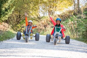Woman and child ride mountain karts on gravel path, smiling and raising arms in excitement. | © Elisabeth Wolkenstein