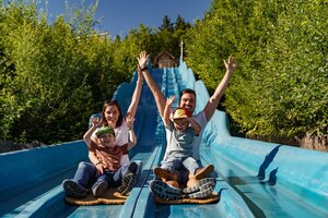 Two adults and two kids slide down a blue wave slide side by side, all laughing and raising their hands. | © Christine Höflehner