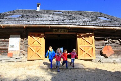 Fünf Kinder rennen lachend aus dem Holzgebäude „Kinderstadl“ bei sonnigem Wetter. | © Katharina Wassler
