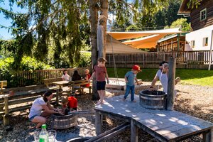 Kinder spielen barfuß mit Wasser und Holzfässern im schattigen Spielplatzbereich am Wilden Berg in Mautern. | © Christine Höflehner