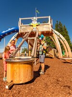 Family plays at Hopsiland Planai with water chute in front, wooden play tower with blue slide and bunny figure in background. | © Christine Höflehner