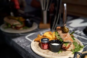 Crispy baguette with bacon, rocket, sauces, pickle and wedges served on a wooden plate in bright sunlight on a black tablecloth. | © Dietmar Körbler