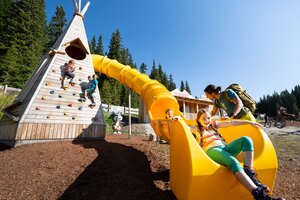 Kids climb a wooden play tower with yellow slide at Hopsiland Planai, one girl slides down smiling, father standing next to her. | © Josh Absenger