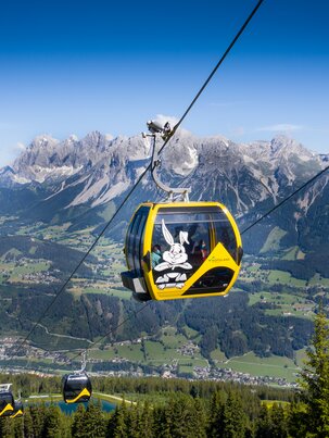 Yellow gondola with Hopsiland bunny above Planai with Dachstein mountains and green Enns valley in bright summer weather. | © Josh Absenger
