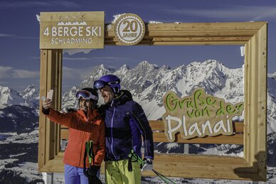 Two skiers take a selfie in front of a wooden frame with snowy mountains. | © Herbert Raffalt