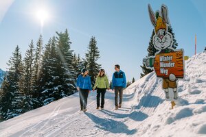 Three hikers walk along the winter hiking trail with Hopsi mascot in the background. | © Christine Höflehner