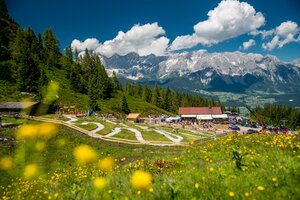Kindergerechte Junior Bike Trails auf der Reiteralm mit Familien, blühender Wiese, Almhütte und Blick auf das Dachsteinmassiv. | © Reiteralm Bergbahnen