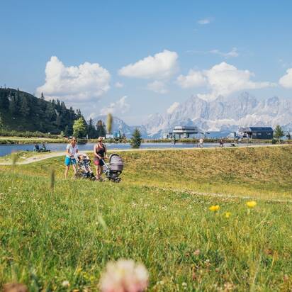 Familien mit Kinderwagen spazieren an einem Bergsee auf der Reiteralm entlang, umgeben von Blumenwiese, Hütten und Bergen. | © Gerald Grünwald