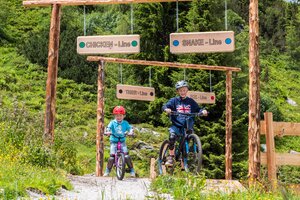 Zwei Kinder fahren mit Mountainbikes in die beschilderten Junior Trails der Reiteralm, umgeben von Blumenwiese und Wald. | © Reiteralm Bergbahnen