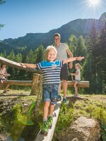 Ein Junge balanciert lachend über einen Holzsteg am Bach, begleitet von Familie beim sonnigen Naturerlebnis auf der Reiteralm. | © Lorenz Masser
