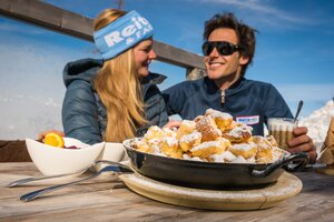 Kaiserschmarrn in a pan on a wooden table, man and woman talking, Reiteralm background. | © Lorenz Masser