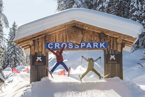 Two skiers jumping at the same time under a wooden ""Crosspark"" sign at Reiteralm. | © Lorenz Masser