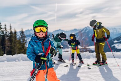 A boy in ski gear stands grinning on the slope and in the background two adults are talking to a child.