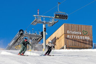 Zwei Skifahrer fahren bei Sonne vor dem Erlebnis Rittisberg und dem Sessellift talwärts.