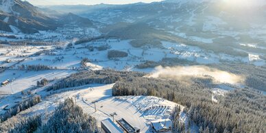Drohnenaufnahme vom verschneiten Rittisberg mit Talstation, Piste und Ennstal im Sonnenlicht.