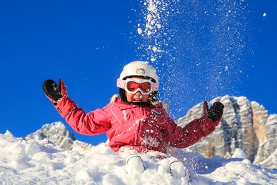 Kind in pinker Skibekleidung spielt lachend im Schnee vor blauer Himmel und Bergen.