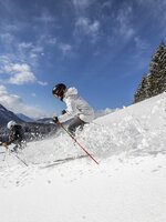 Two skiers descend through deep powder snow in sunshine near forest edge | © filzmoos.ski