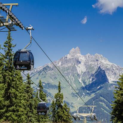 Papagenobahn cable cars schweben über Nadelwald in Filzmoos, dahinter ragt ein markantes Felsmassiv auf. | © filzmoos.ski