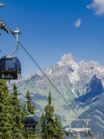 Papagenobahn cable cars schweben über Nadelwald in Filzmoos, dahinter ragt ein markantes Felsmassiv auf. | © filzmoos.ski