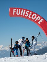 A family with two children stands under a red half arch on the Funslope and each holds a ski high in the air