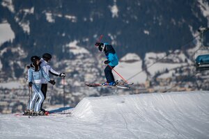 Skier in blue gear jumps at Beginnerpark with snowy backdrop.