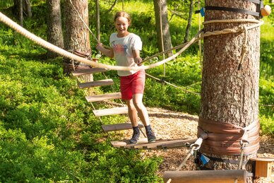 Girl crosses hanging bridge in forest rope course on a sunny day. | © Shuttleberg