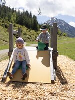 Two small children play on a low wooden slide with green mountains behind. | © Shuttleberg