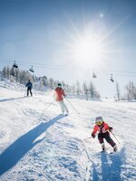 Child and adults skiing on a sunny slope, with chairlift and snowy trees in the background | © Shuttleberg